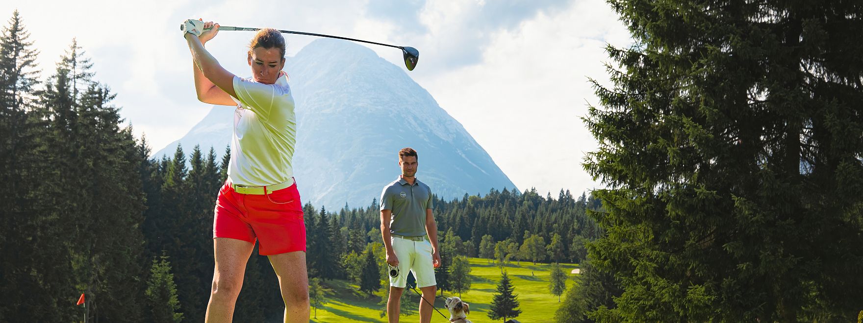 Golfen im GC Wildmoos im Sommer_Golferin bei Abschlag vor Hohe Munde_Mann mit Hund im Hintergrund.jpg